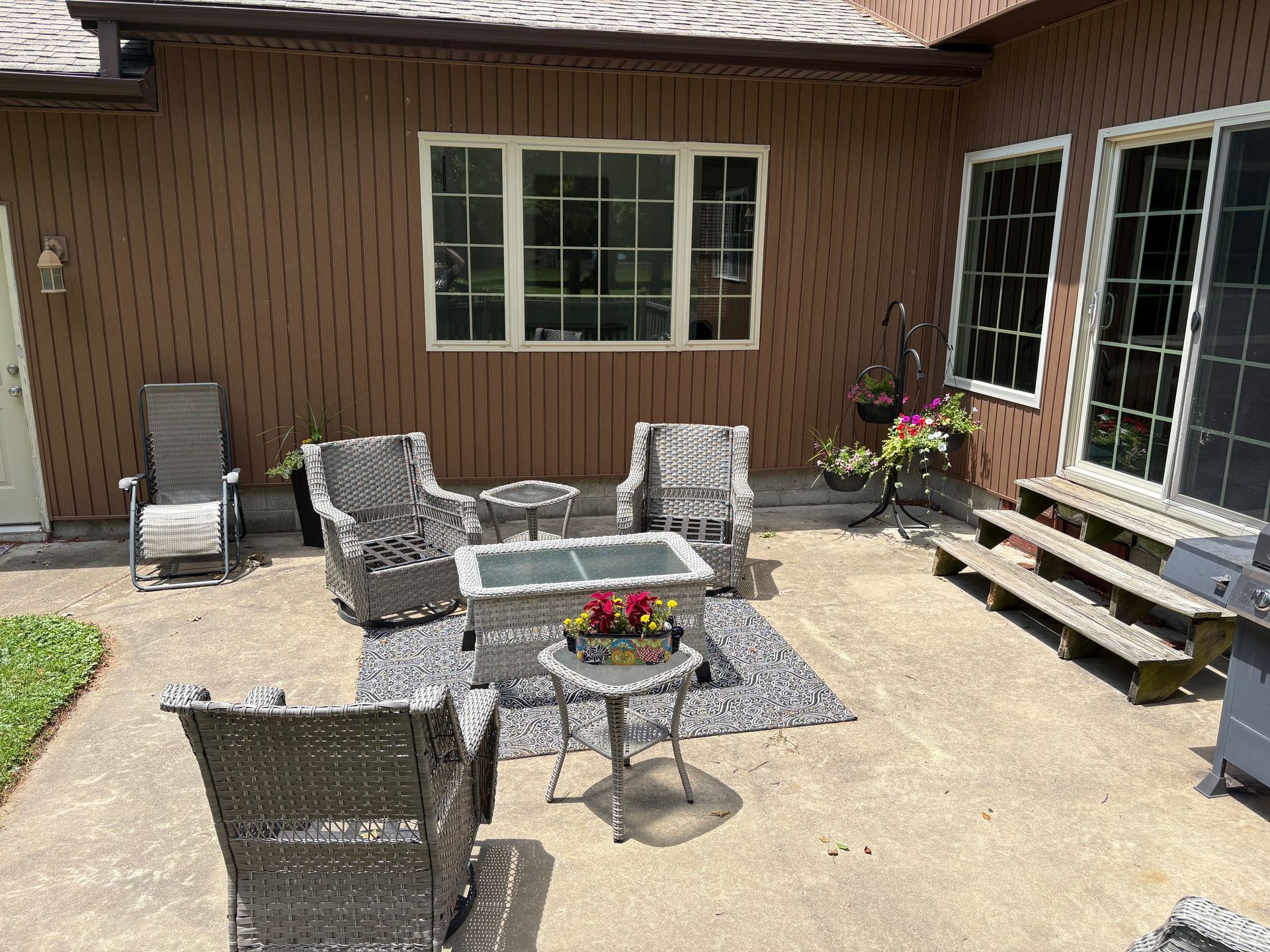 Patio seating area with wicker chairs, small tables, flower pots, and steps by a house wall