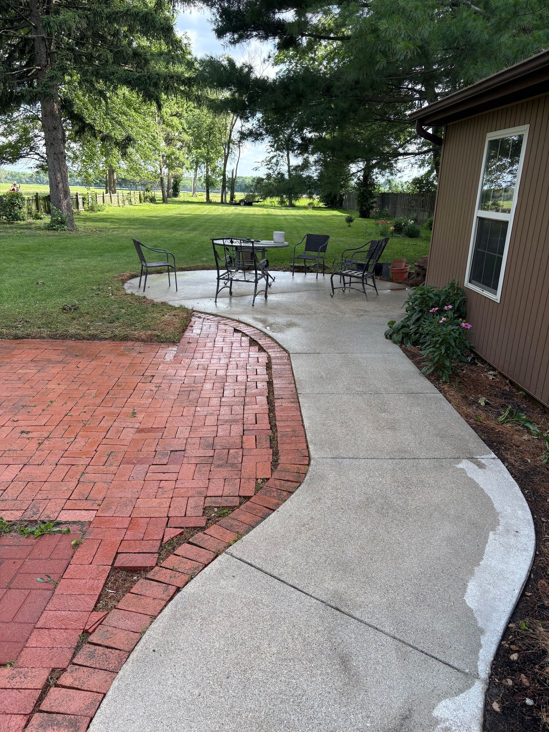Brick and concrete patio path beside a house, leading to outdoor chairs on a grassy yard.