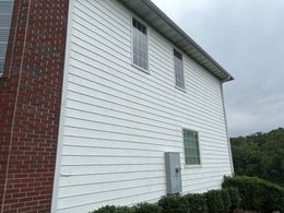 White-sided building with boarded windows, a gray door, and red brick corner under cloudy sky