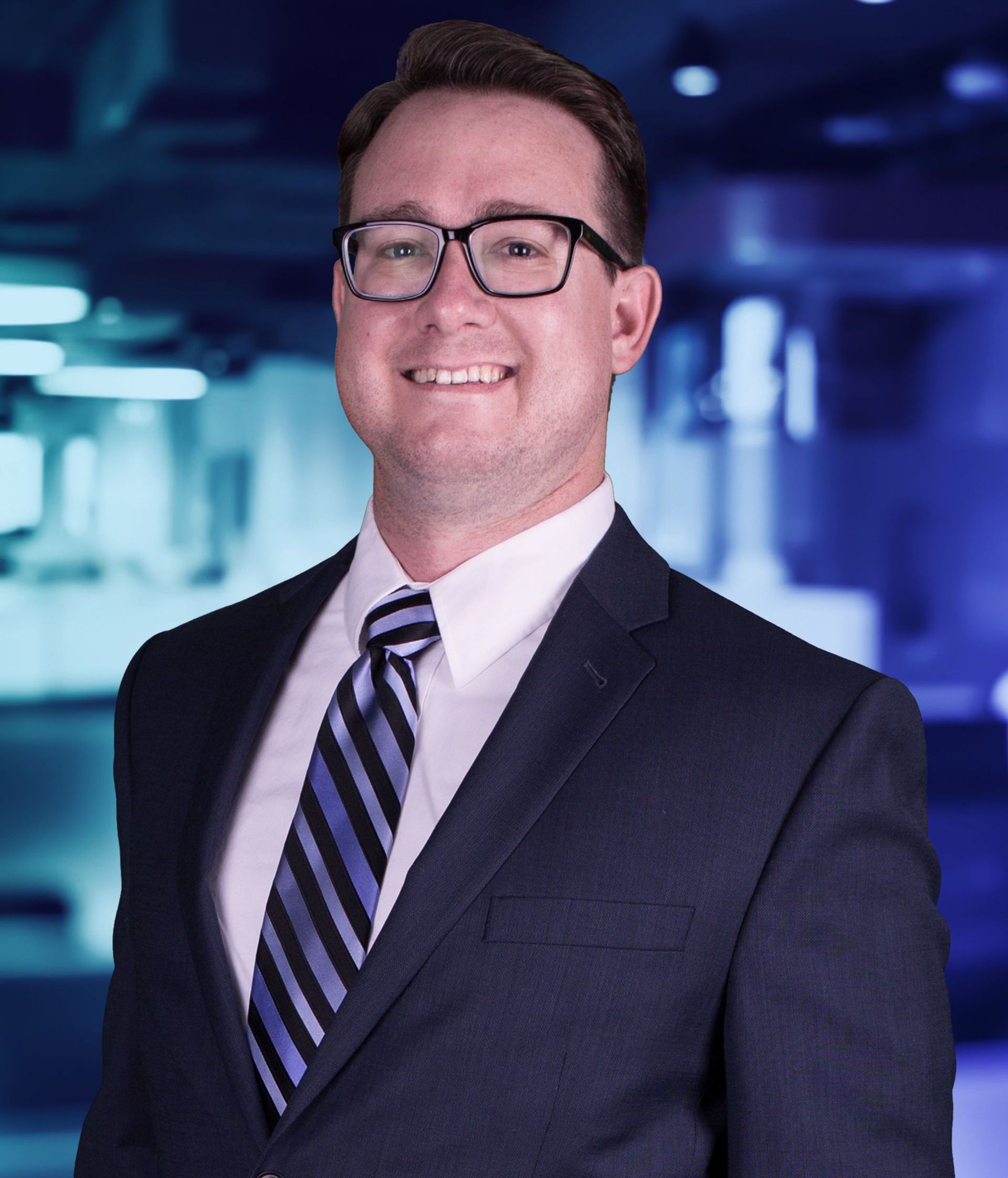 Man in suit and glasses smiles in a professional setting. He wears a tie with blue and black stripes.