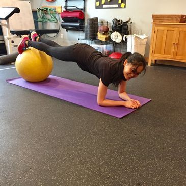 A woman is doing a plank on a purple mat next to a yellow ball