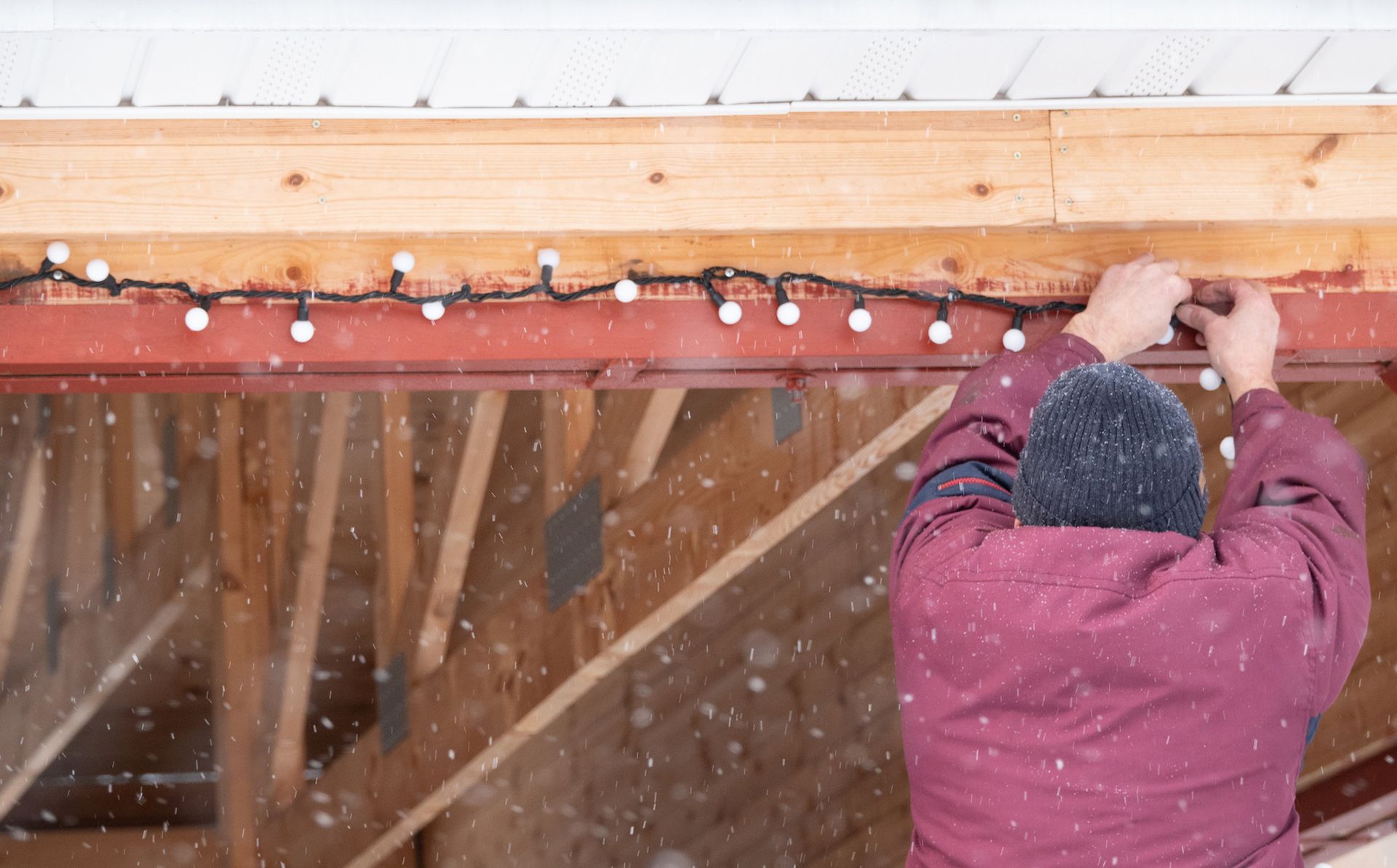 Person hangs white Christmas lights along a red metal beam on a wooden structure, snowing.