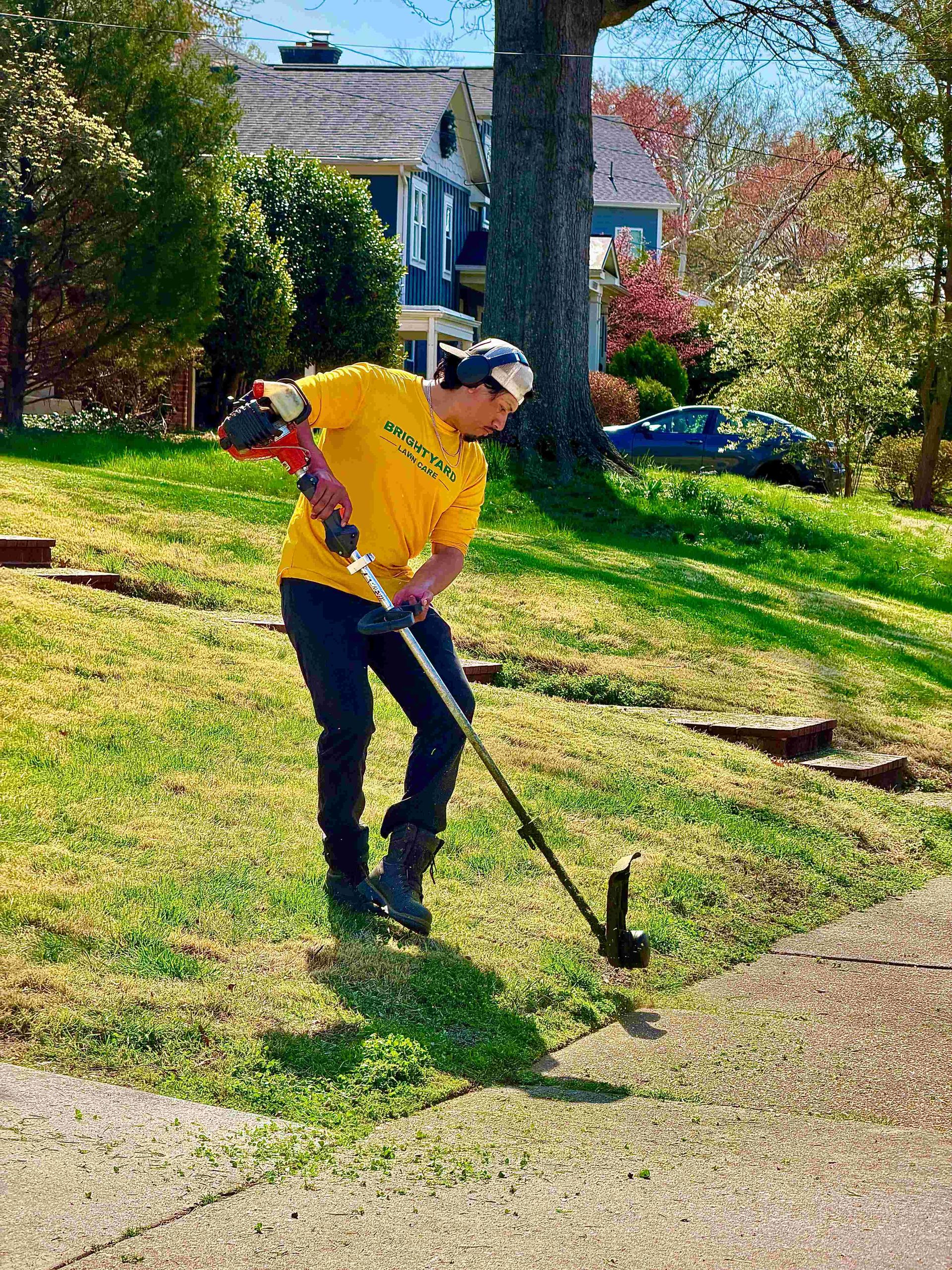 A person in a yellow shirt trims the edge of a grassy lawn along a sidewalk with a string trimmer.