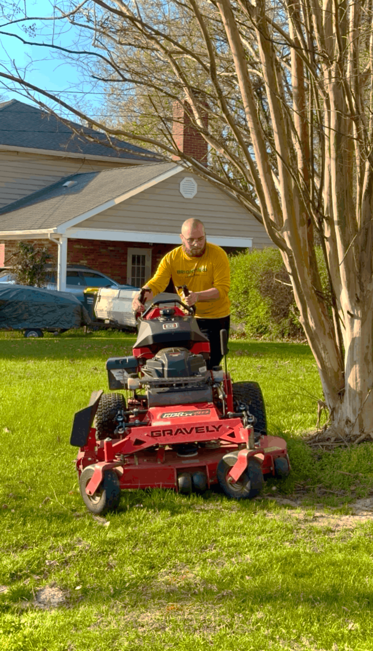 A person in a yellow shirt stands on a red zero-turn mower on a grassy lawn next to a brick building.