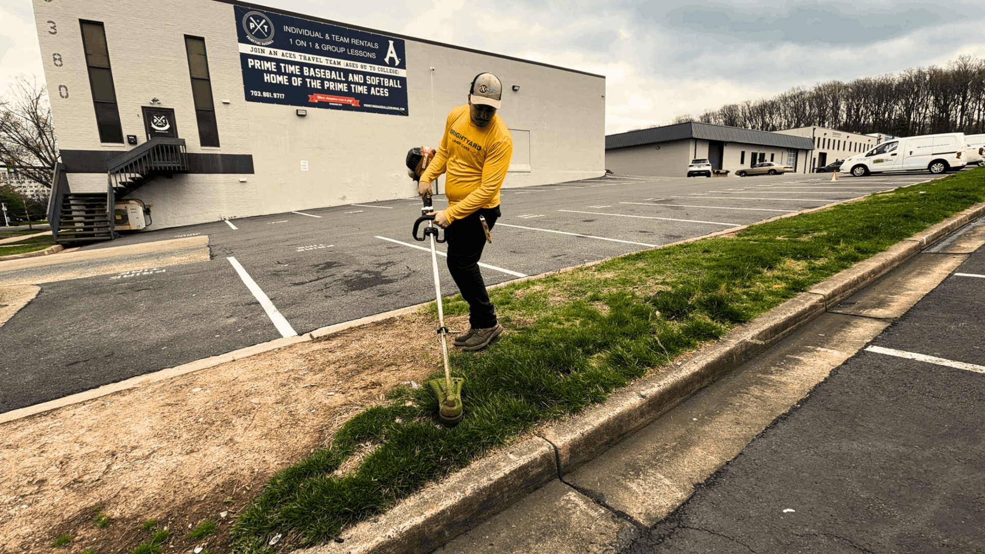 A worker in a yellow shirt uses a string trimmer to trim grass along a concrete curb in a parking lot.