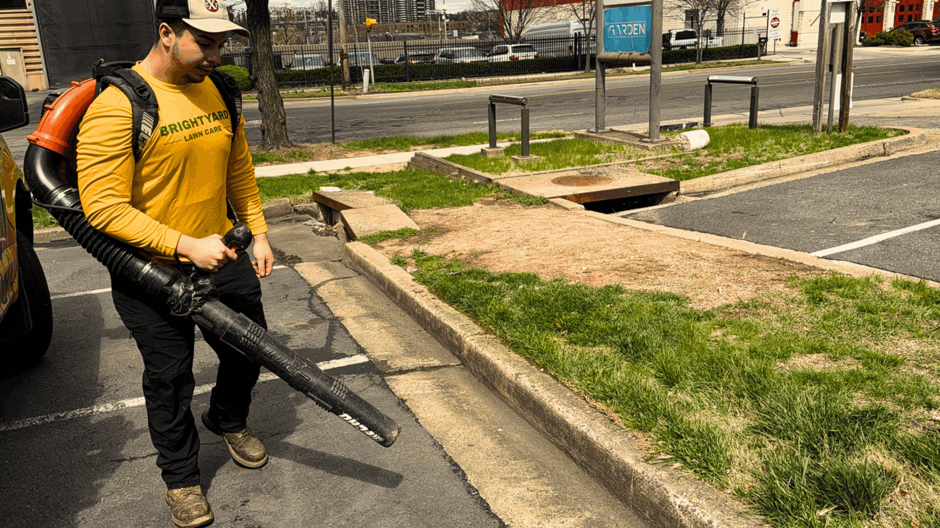 A person in a yellow shirt operates a backpack leaf blower on a paved lot beside a curb and a grassy area.