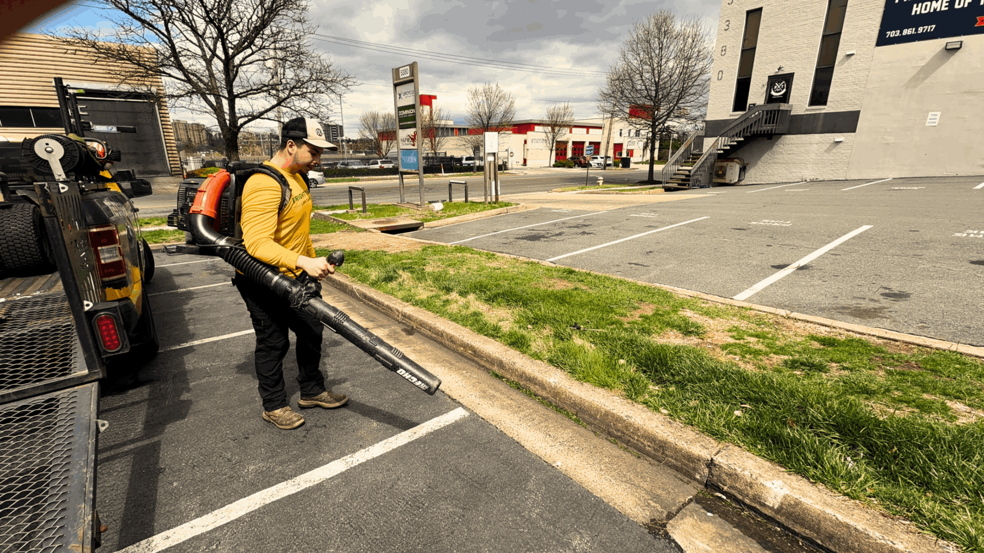 A worker in a yellow long-sleeve shirt uses a backpack leaf blower to clear debris along a parking lot curb.