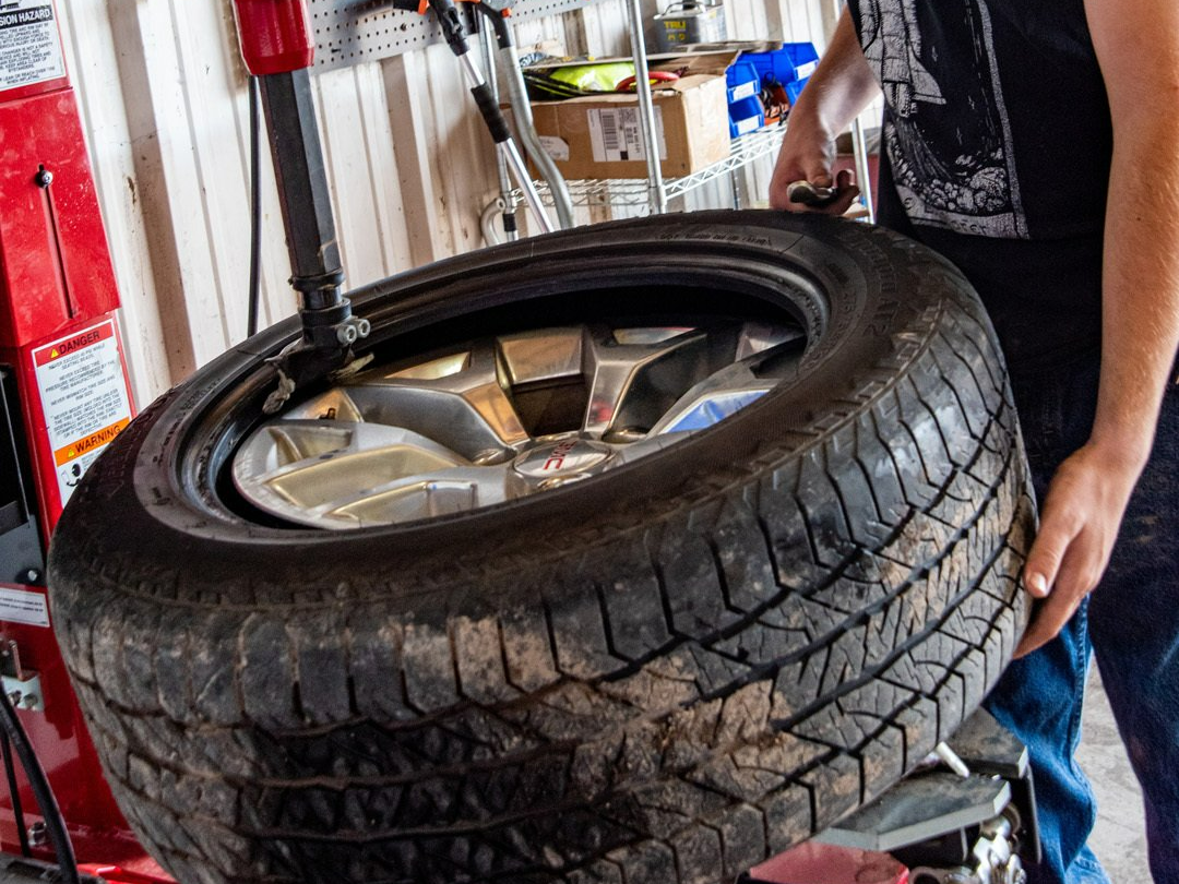 Large black tire on a rim being handled in a garage by a person in jeans and a black shirt