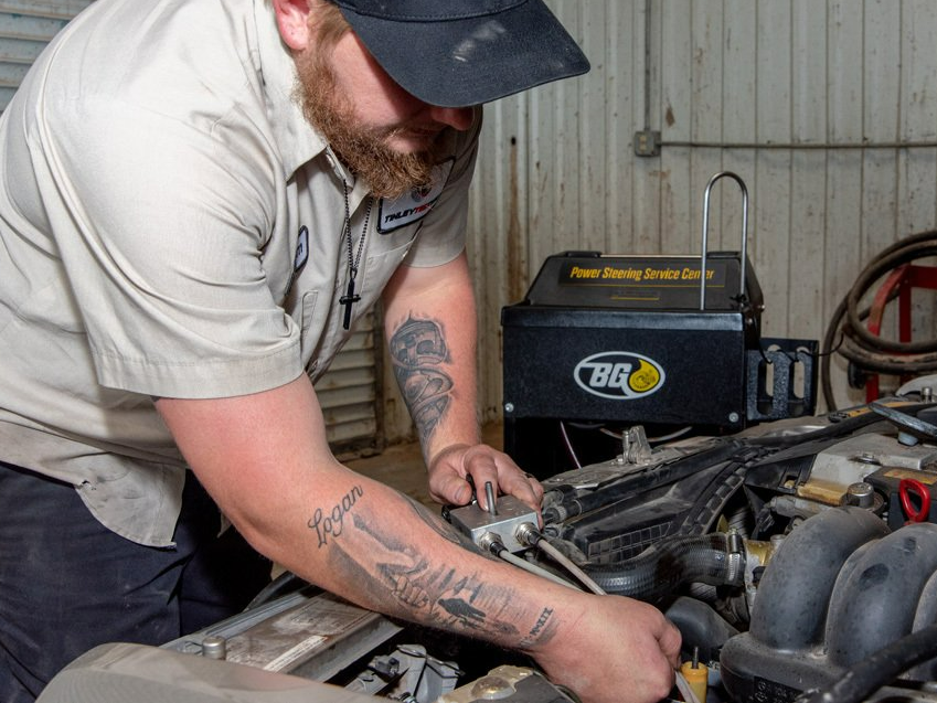 Mechanic working under an open hood in a garage, inspecting engine components with tools.
