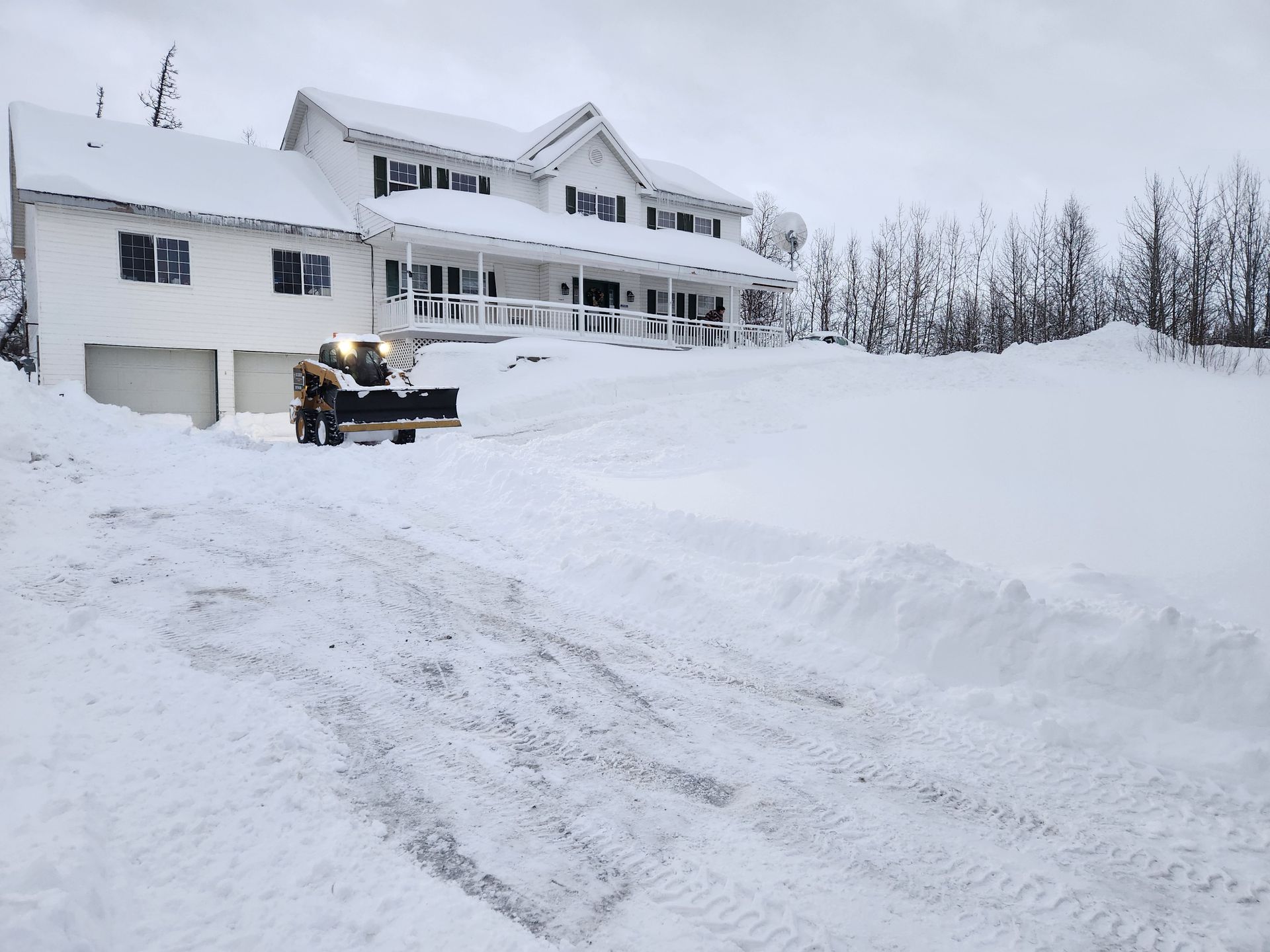 Snow-covered house with a lit snowblower clearing a driveway after heavy snowfall