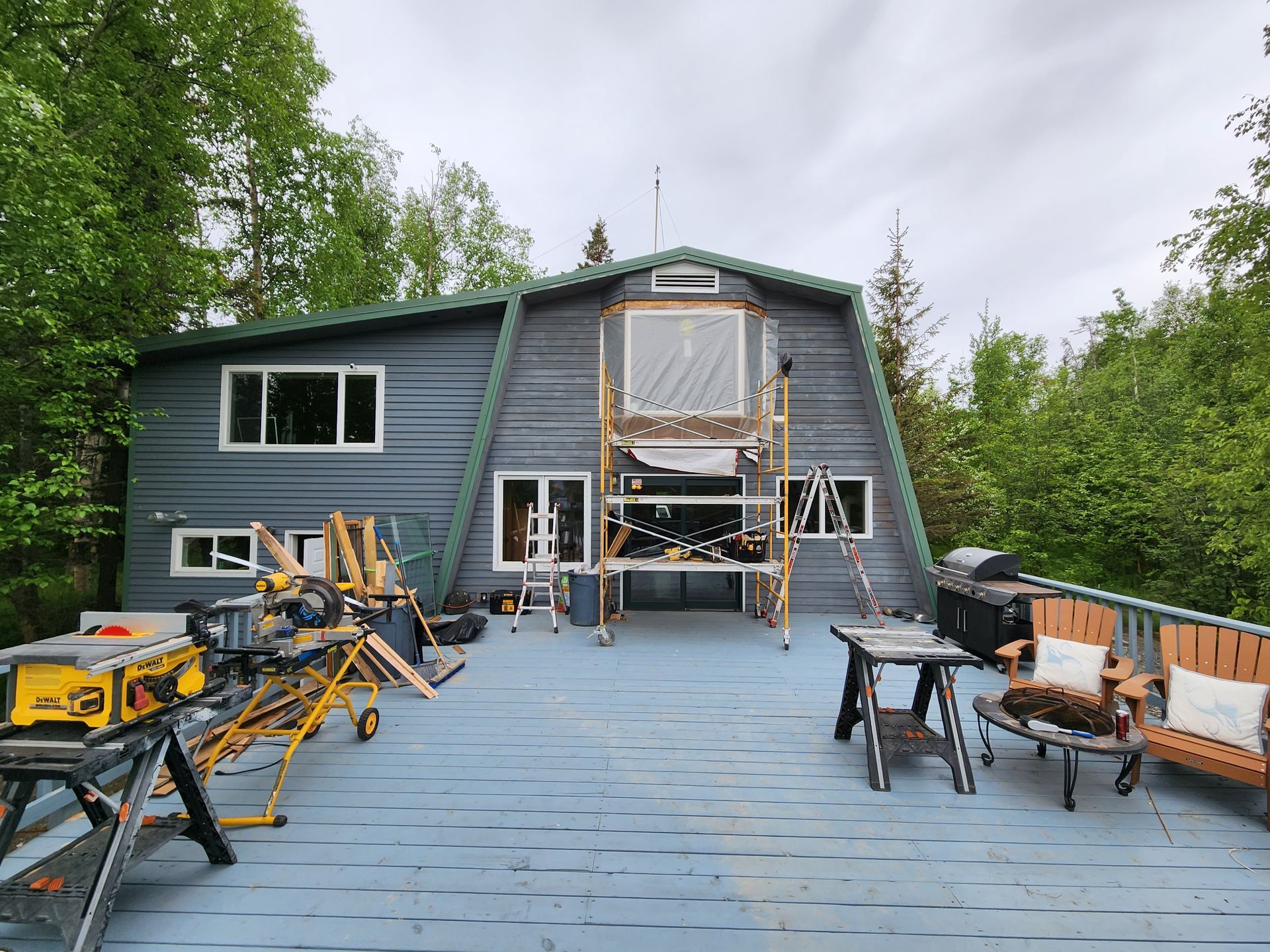 Partially renovated blue house deck with ladders, tools, and outdoor seating surrounded by trees