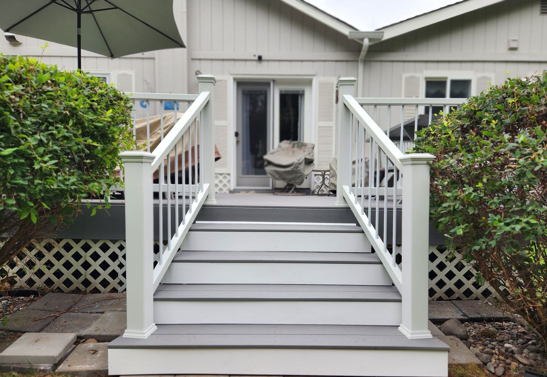 White stairs leading to a patio with railing, a chair, and a large umbrella in front of a house