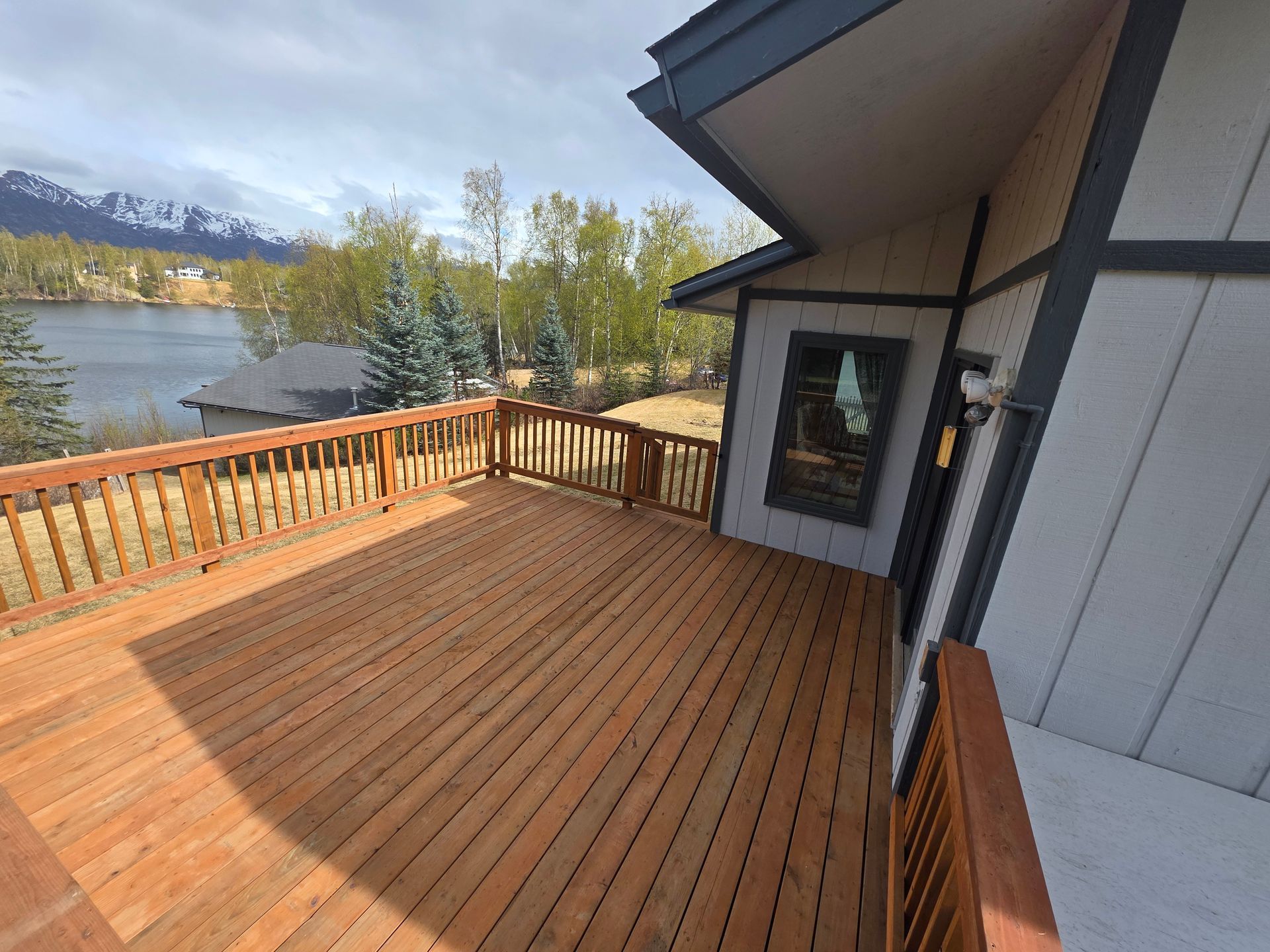 Wooden deck beside a lakeside house, with mountains and trees in the background.