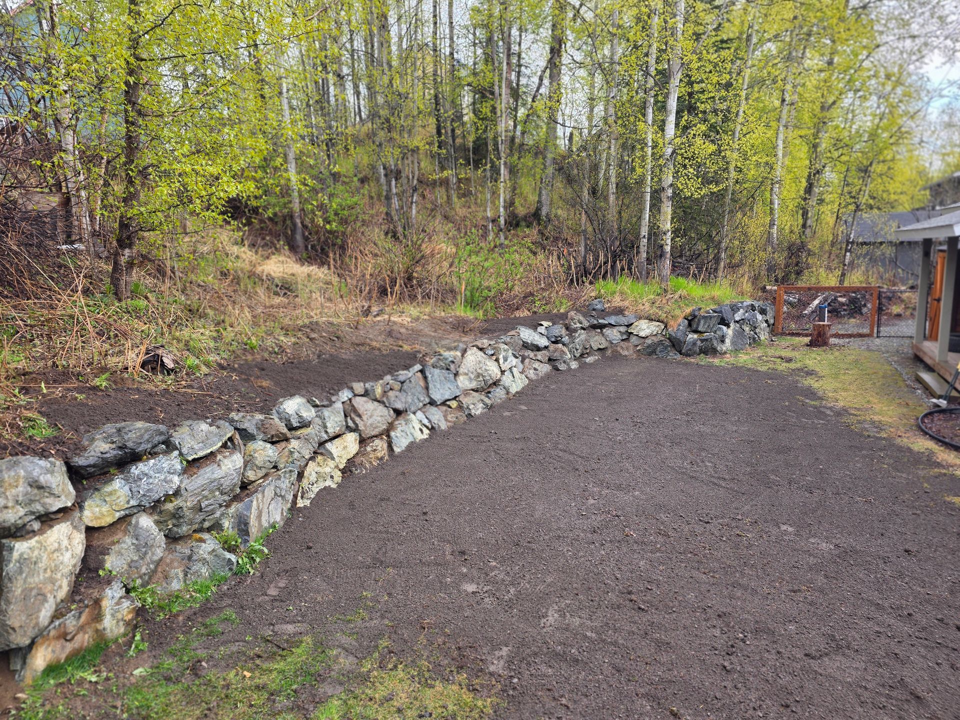 Rock-lined drainage swale beside wet asphalt, with a house and trees in the background