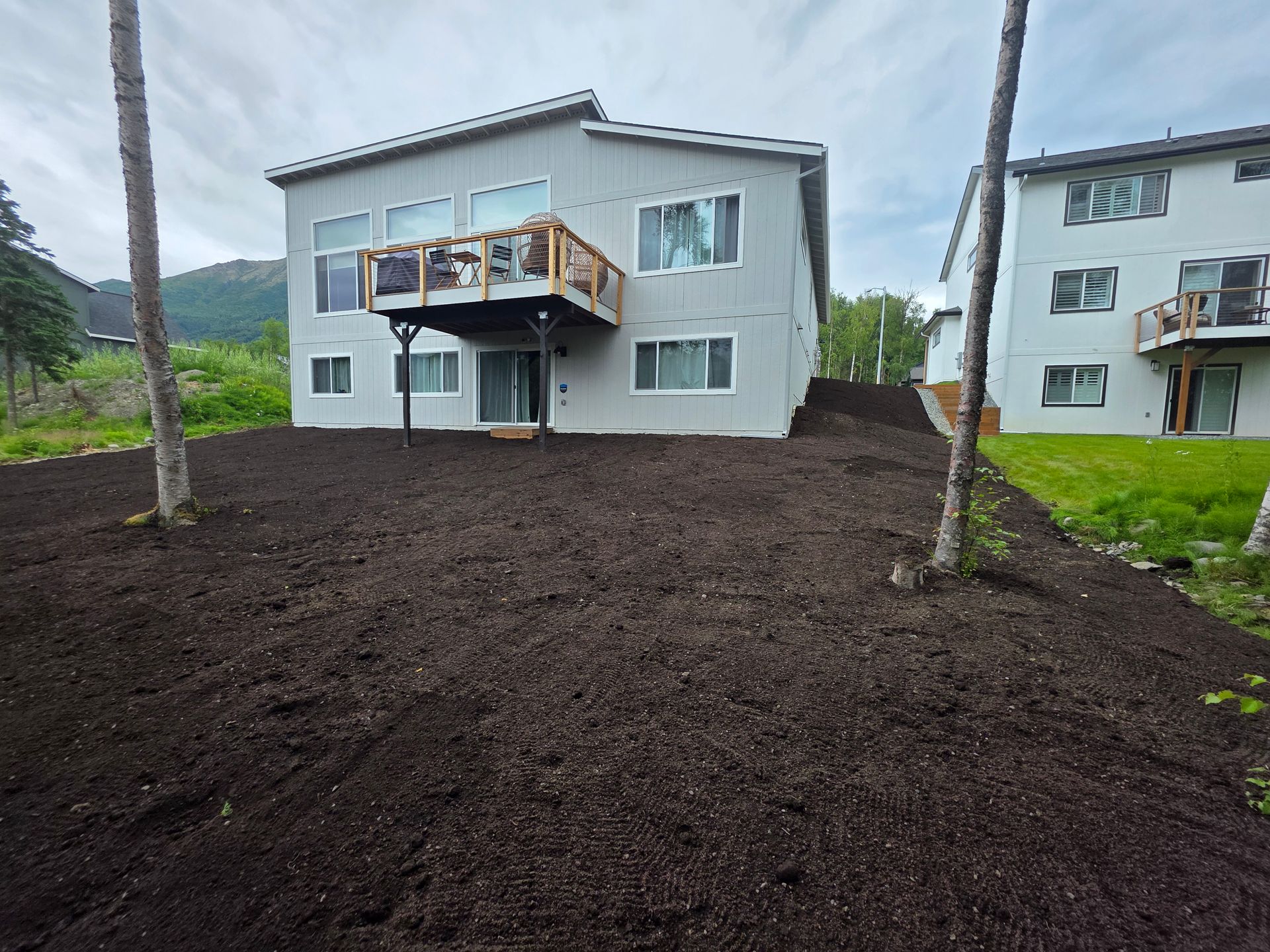 Two-story apartment building with a deck, surrounded by dark soil and palm trees on a cloudy day