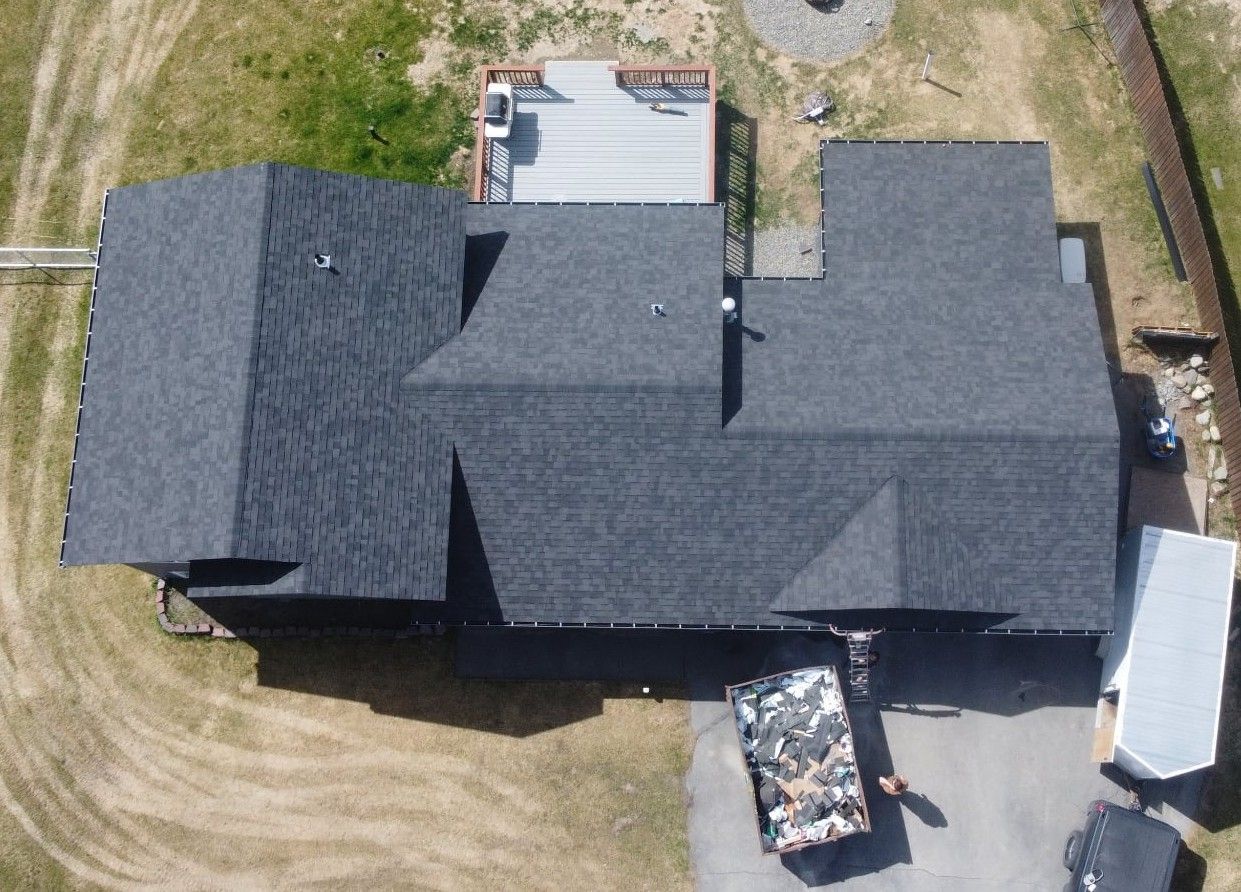 Aerial view of a dark shingle-roof house with a deck, driveway, and trash bin outdoors