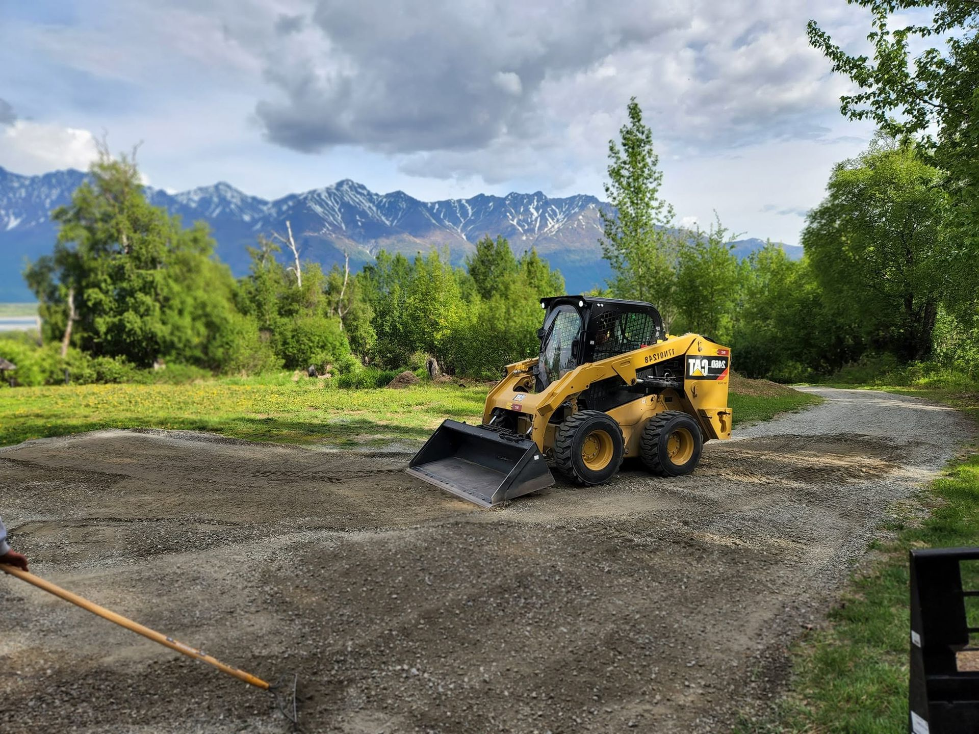 Yellow skid steer on gravel road with mountain backdrop and green trees