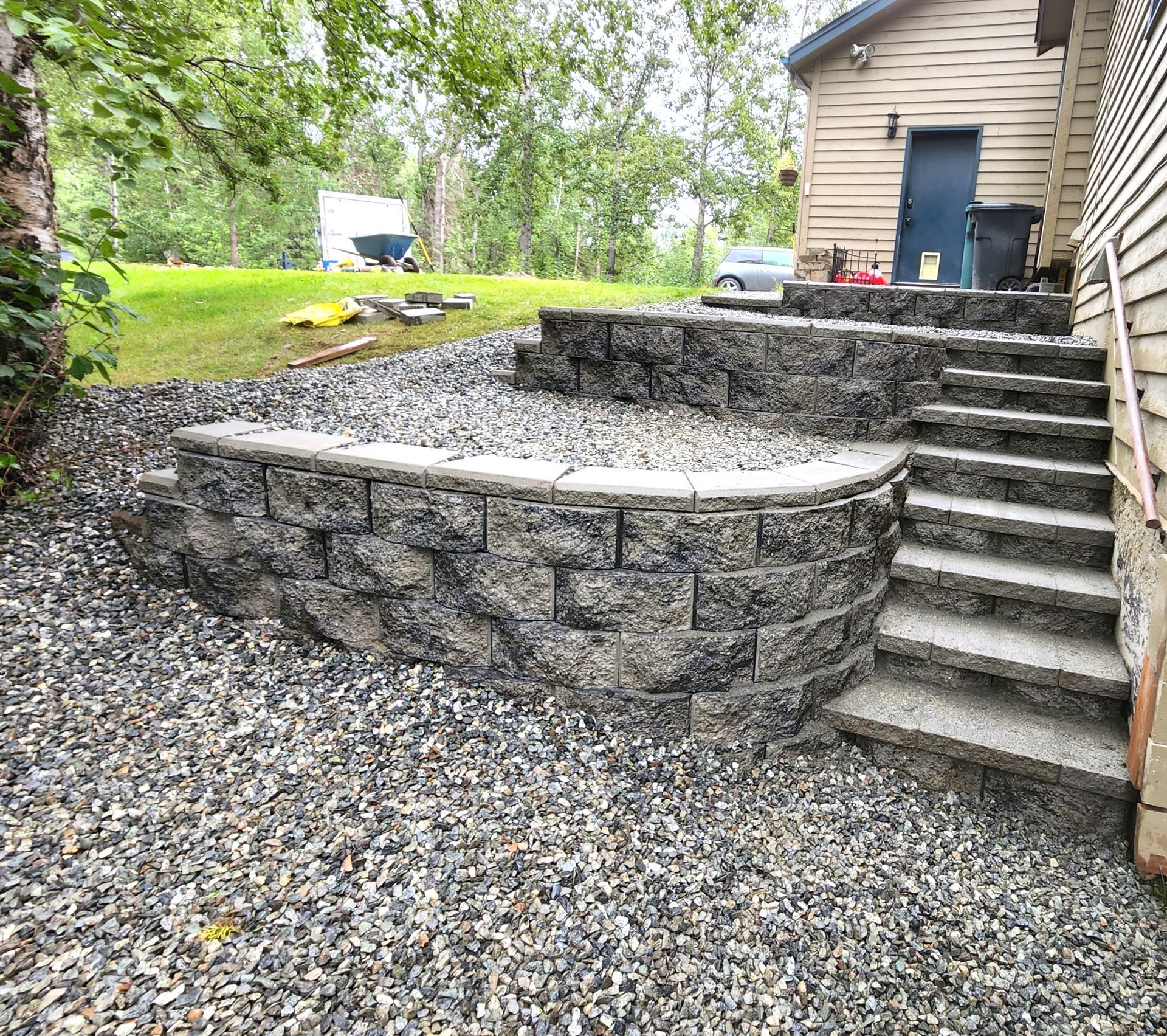 Stone patio and retaining wall with curved seating area beside house and stairs, surrounded by gravel and grass