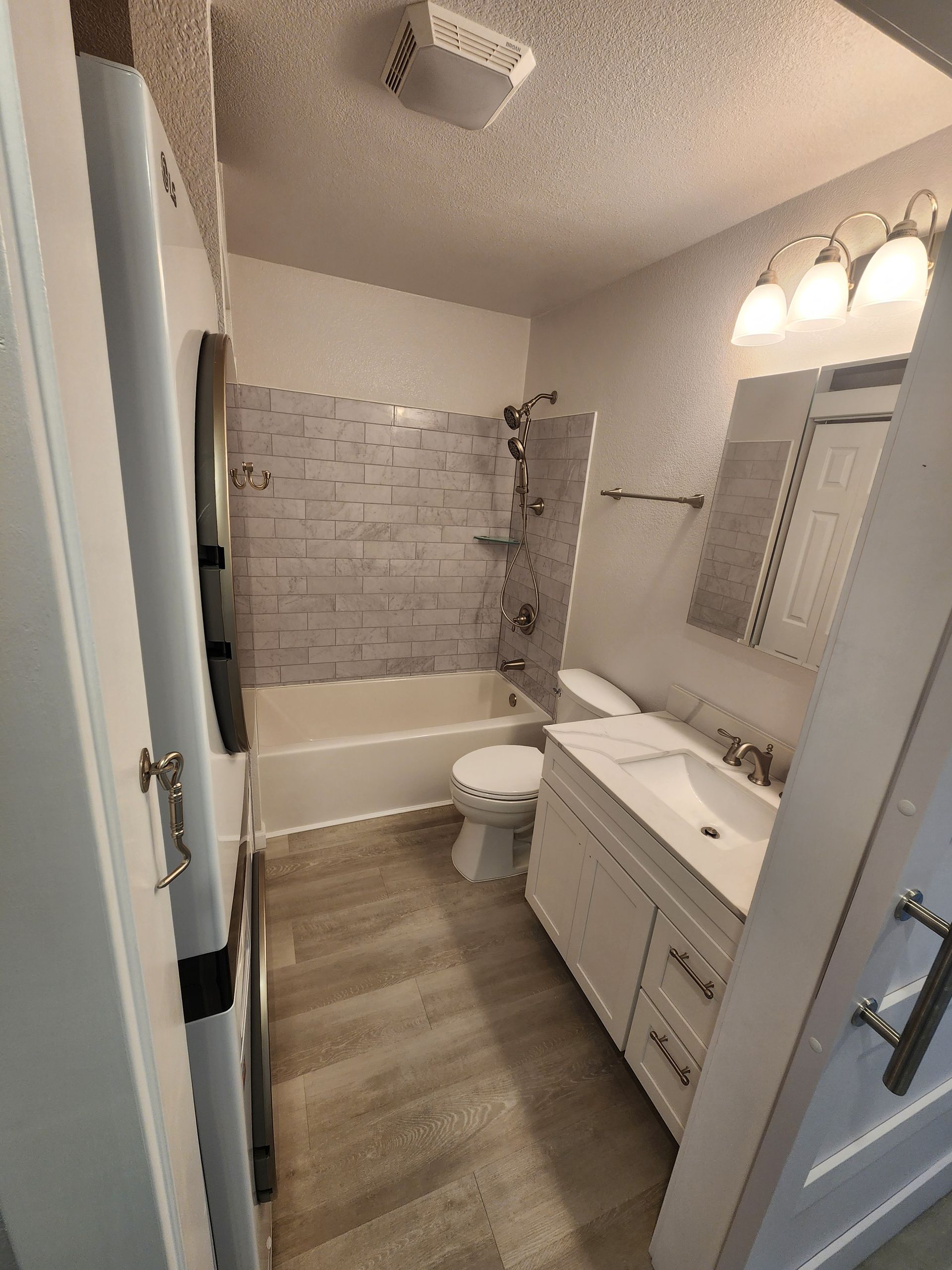 Small bathroom with bathtub, toilet, vanity, mirror, and wood-look floor.