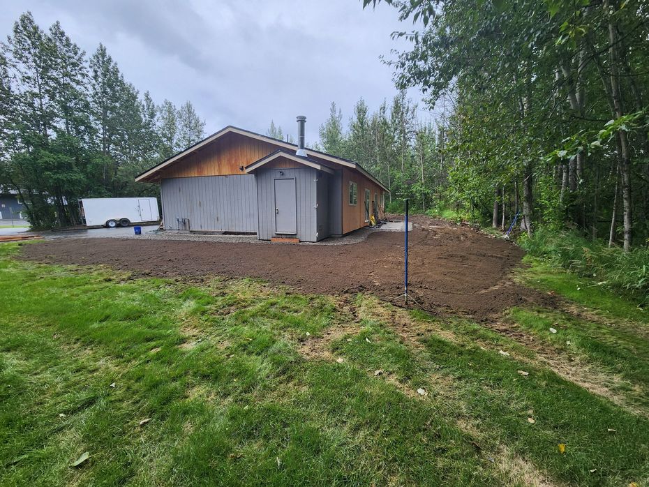 Yellow skid steer dumping dark soil in a residential driveway, with houses in the background