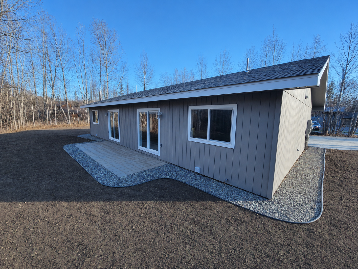 Small beige house with dark roof and white-trimmed windows on a gravel lot under a clear blue sky