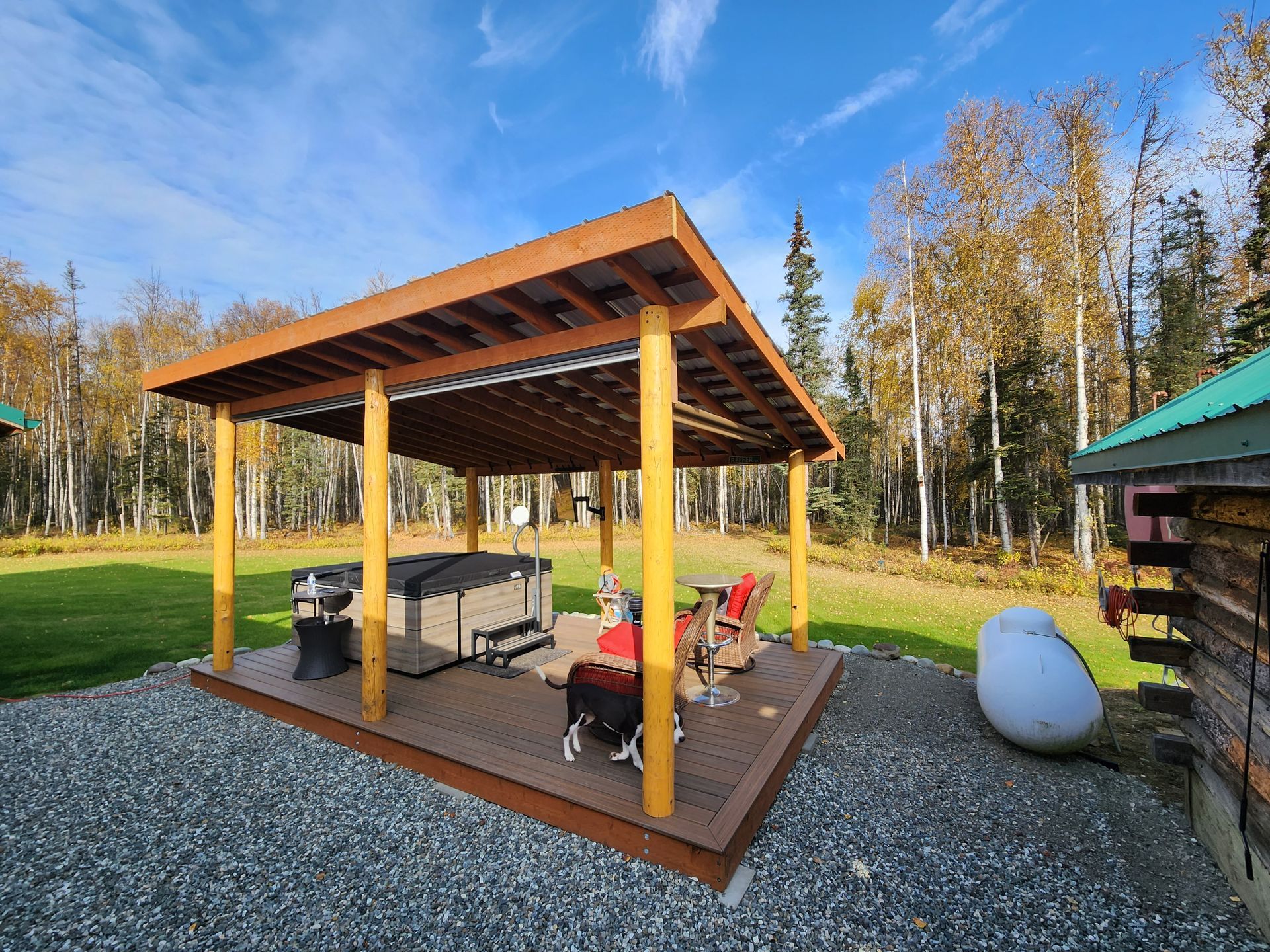 Wooden pergola with picnic table and grill on a gravel patio in a sunny yard