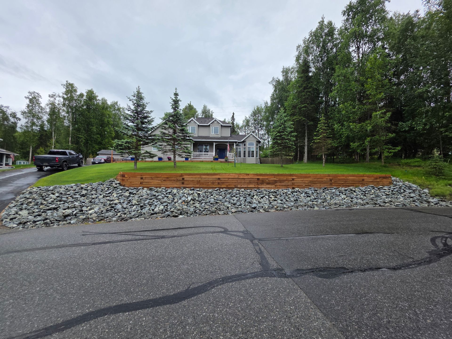 Driveway leading to a large house with trees, lawn, and a raised wooden planter in the foreground