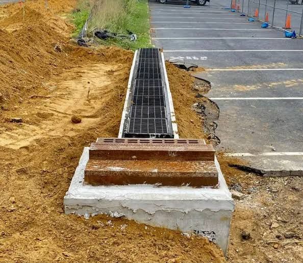 Roadside storm drain inlet with metal grating beside a paved parking lot and exposed soil.