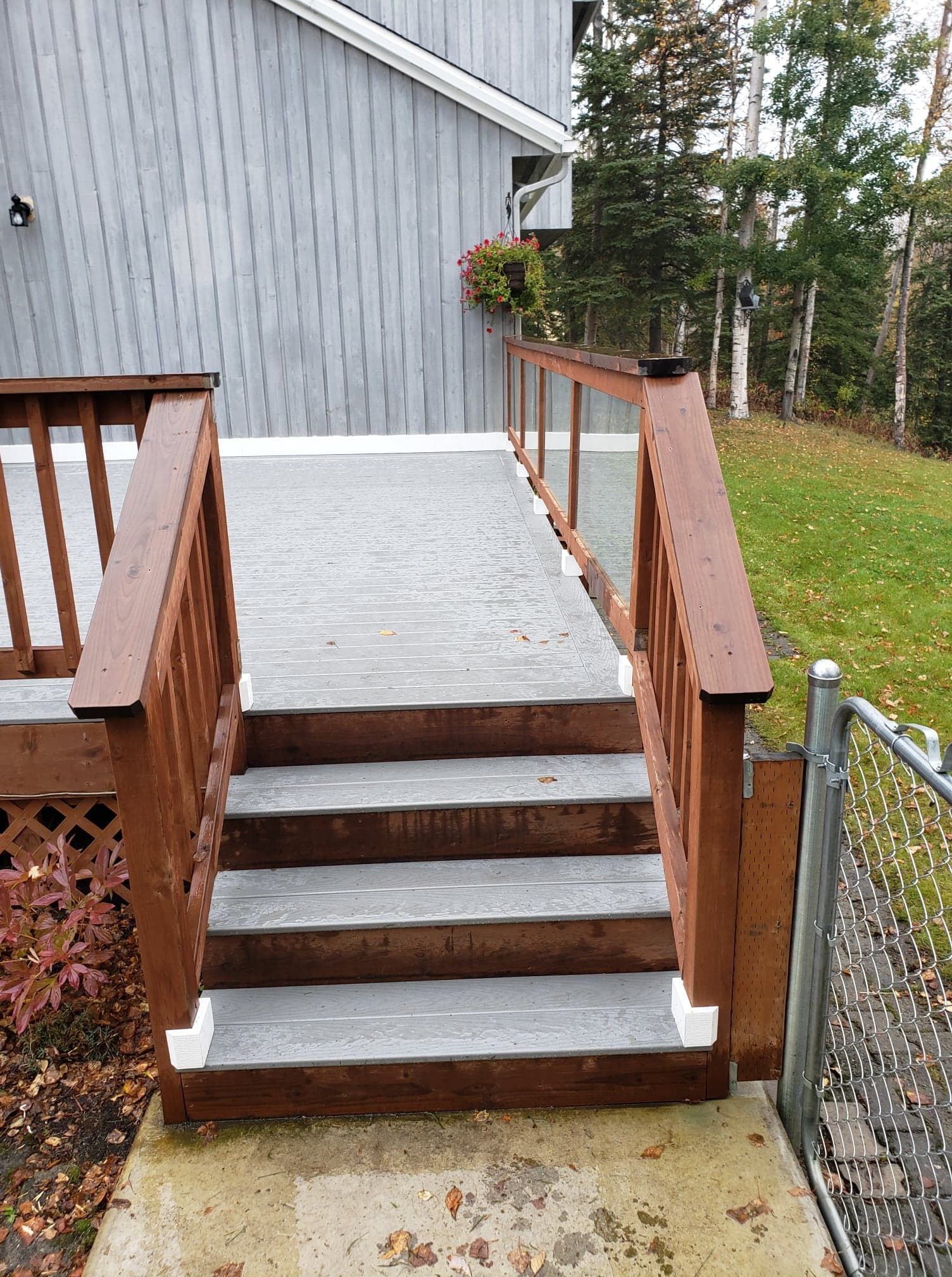 Wooden deck stairs with white treads beside a gray house and chain-link fence, leading to a small patio.