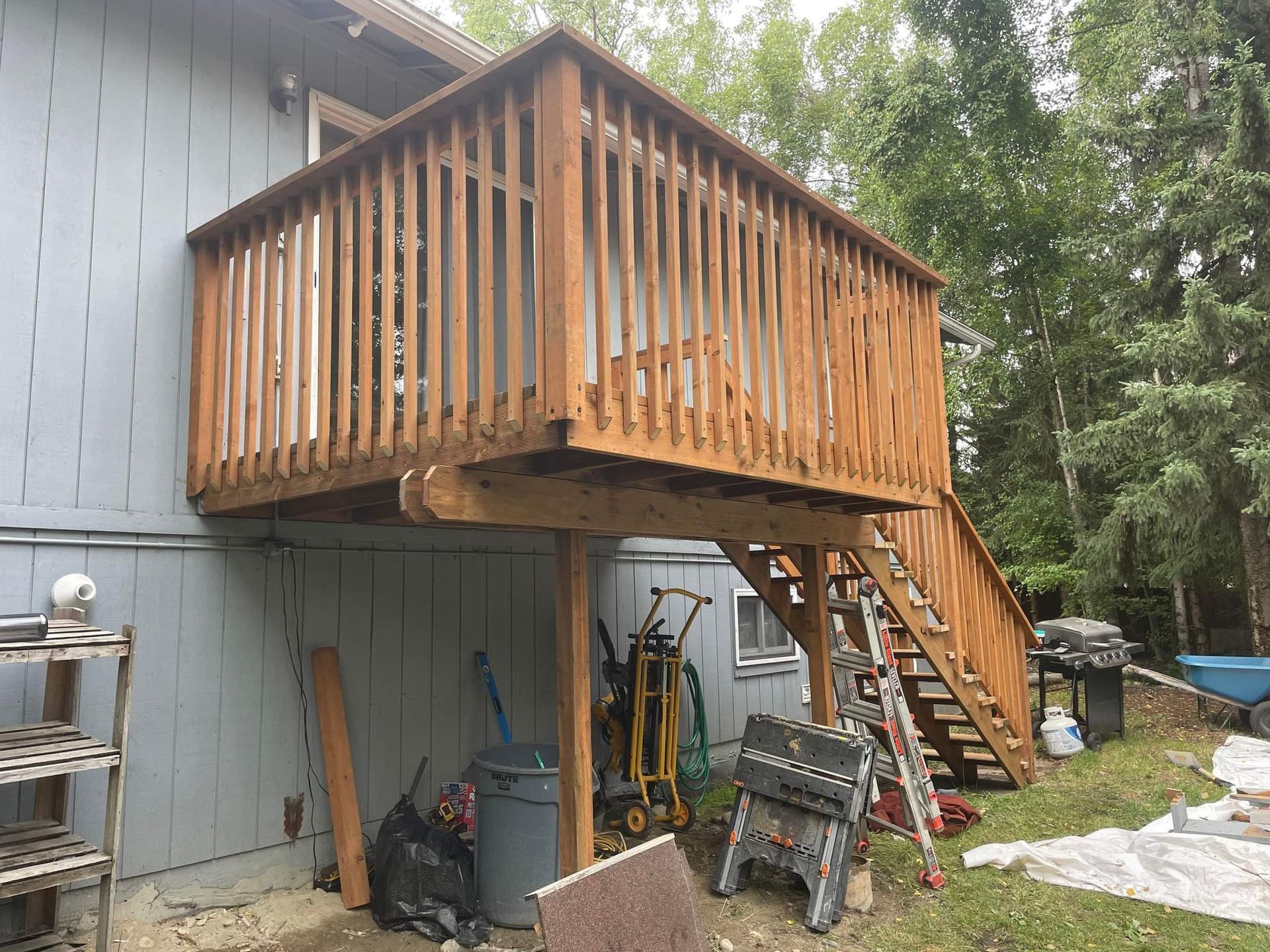 Wooden elevated deck with stairs attached to a gray house, with tools and yard items below.