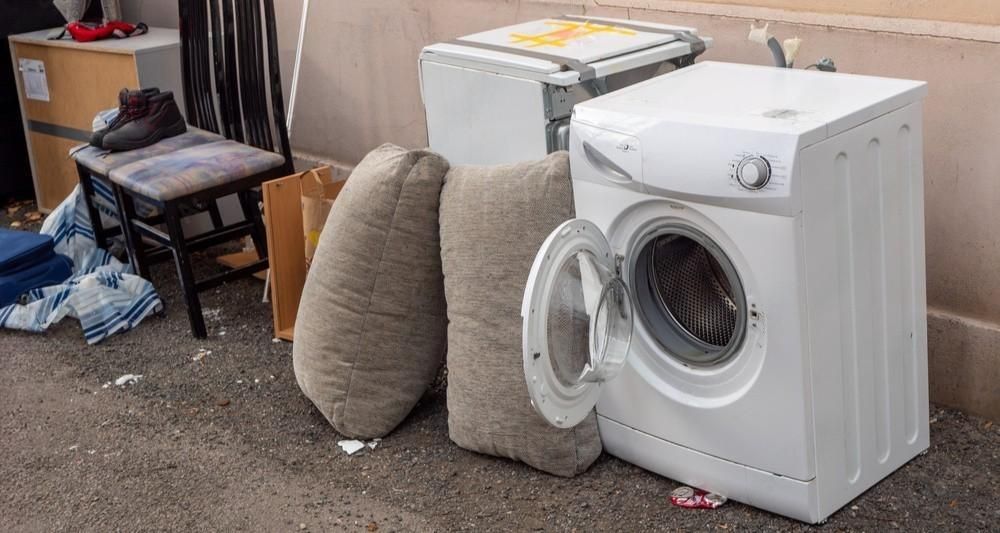 White front-loading washing machine beside two beige cushions and household items in a cluttered room