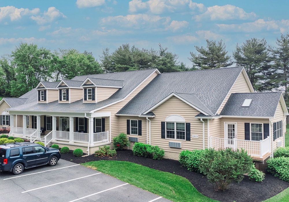 Large beige house with gray roof, white trim, and front porch beside a parking lot and car.