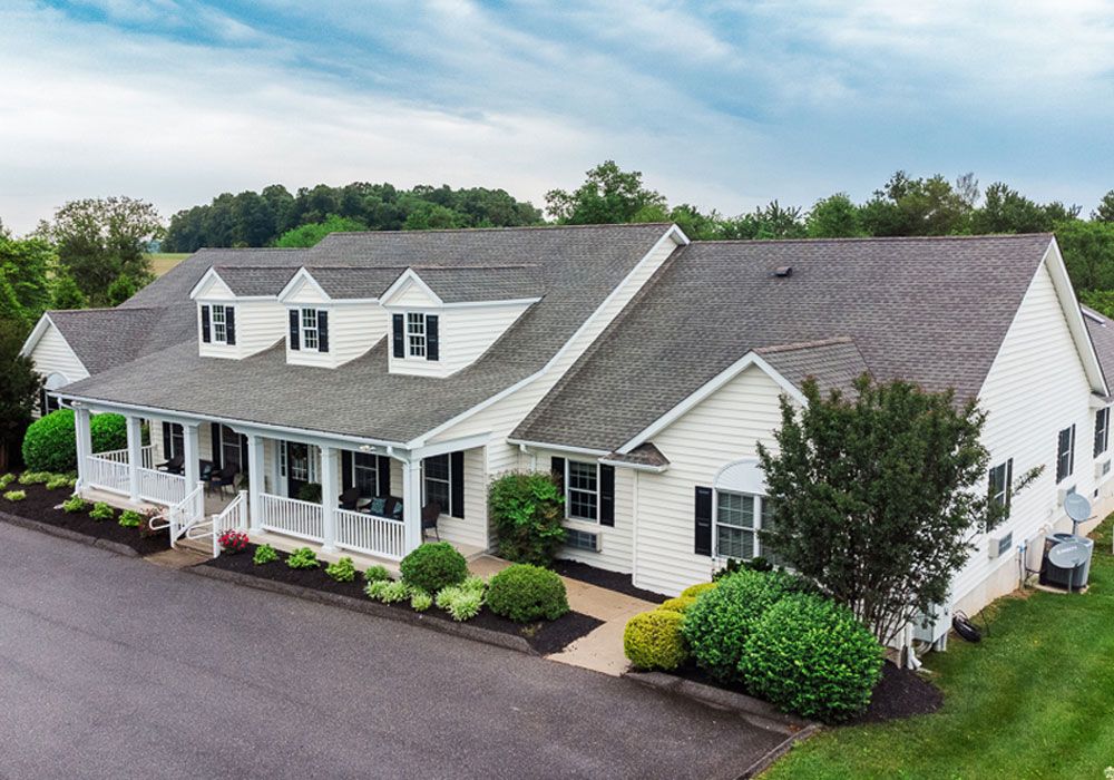 White house with porch and dormer windows, surrounded by landscaping and trees under a cloudy sky.