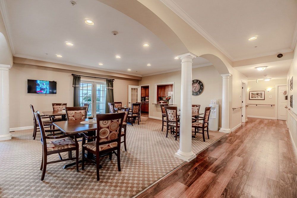 Bright dining room with wooden tables and chairs, white columns, recessed lighting, and a hallway with wood flooring.