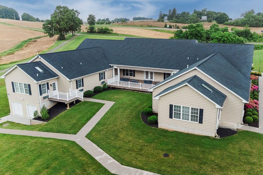Aerial view of a beige house with a dark roof, wraparound porch, and landscaped lawn in a rural setting