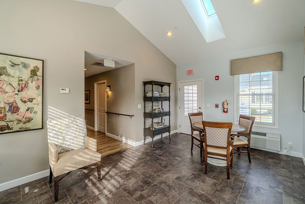 Bright dining area with round table, wooden chairs, gray walls, skylight, and shelving near an open hallway
