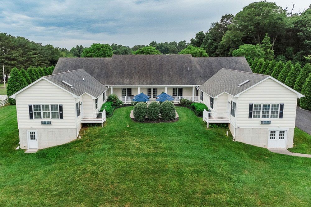 Aerial view of a large white house with a central wing and green lawn, surrounded by trees under cloudy skies