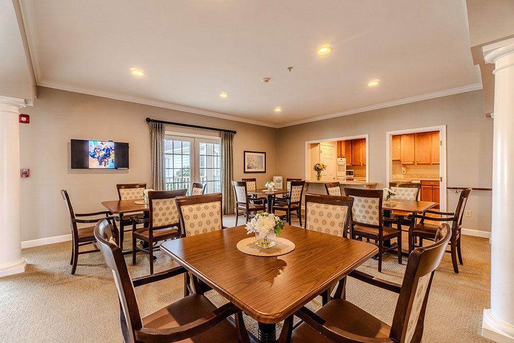 Spacious dining room with wooden tables, chairs, recessed lights, and a view into a kitchen area.