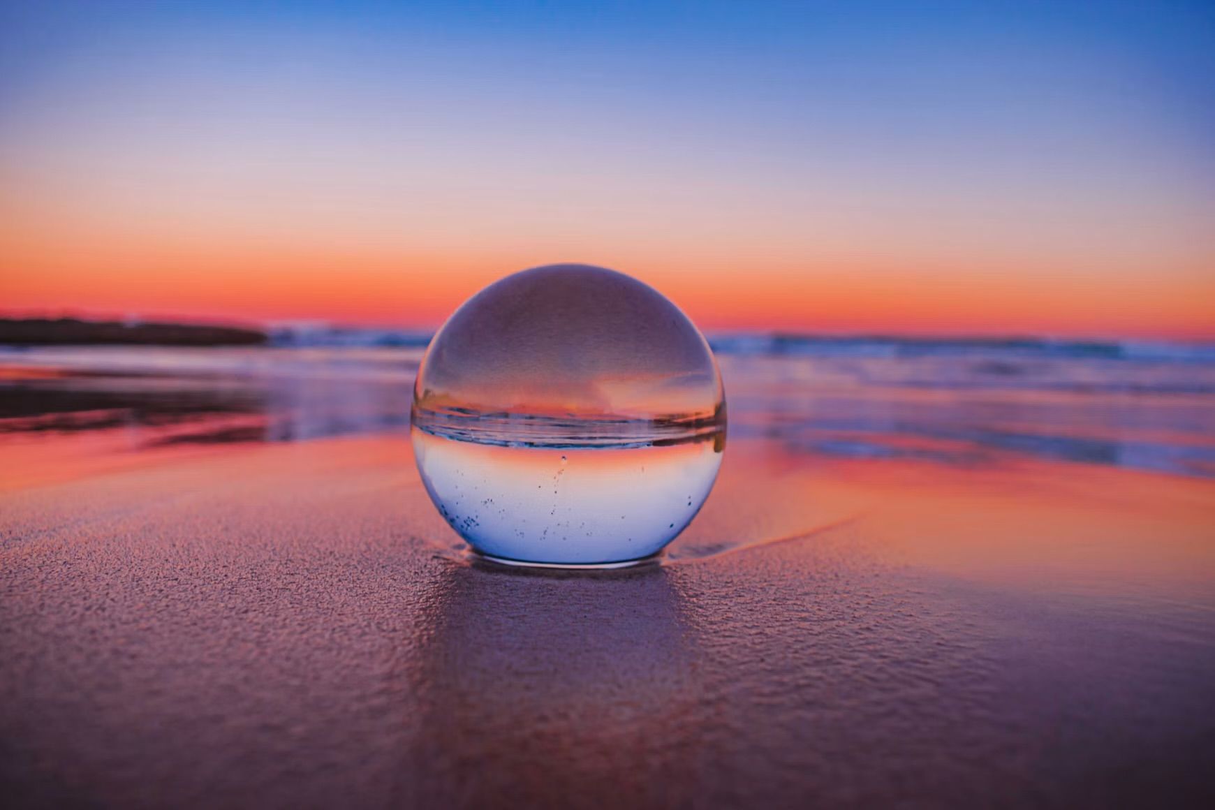 Glass sphere on a beach at sunset, reflecting pink and blue sky over wet sand.