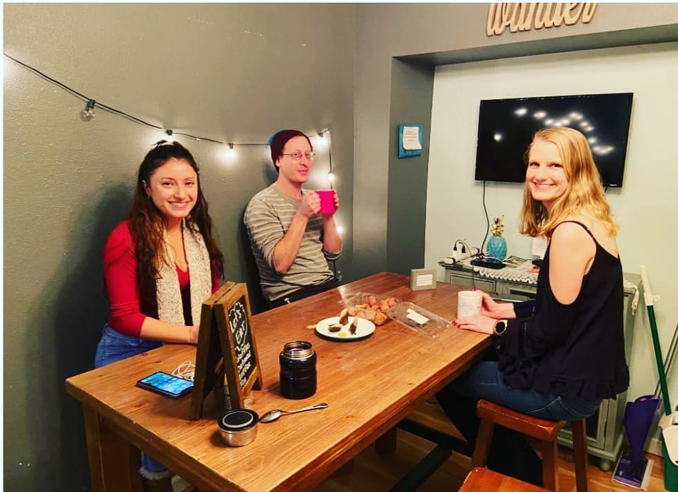 Three people seated at a wooden table, drinking beverages. String lights and TV on the wall.