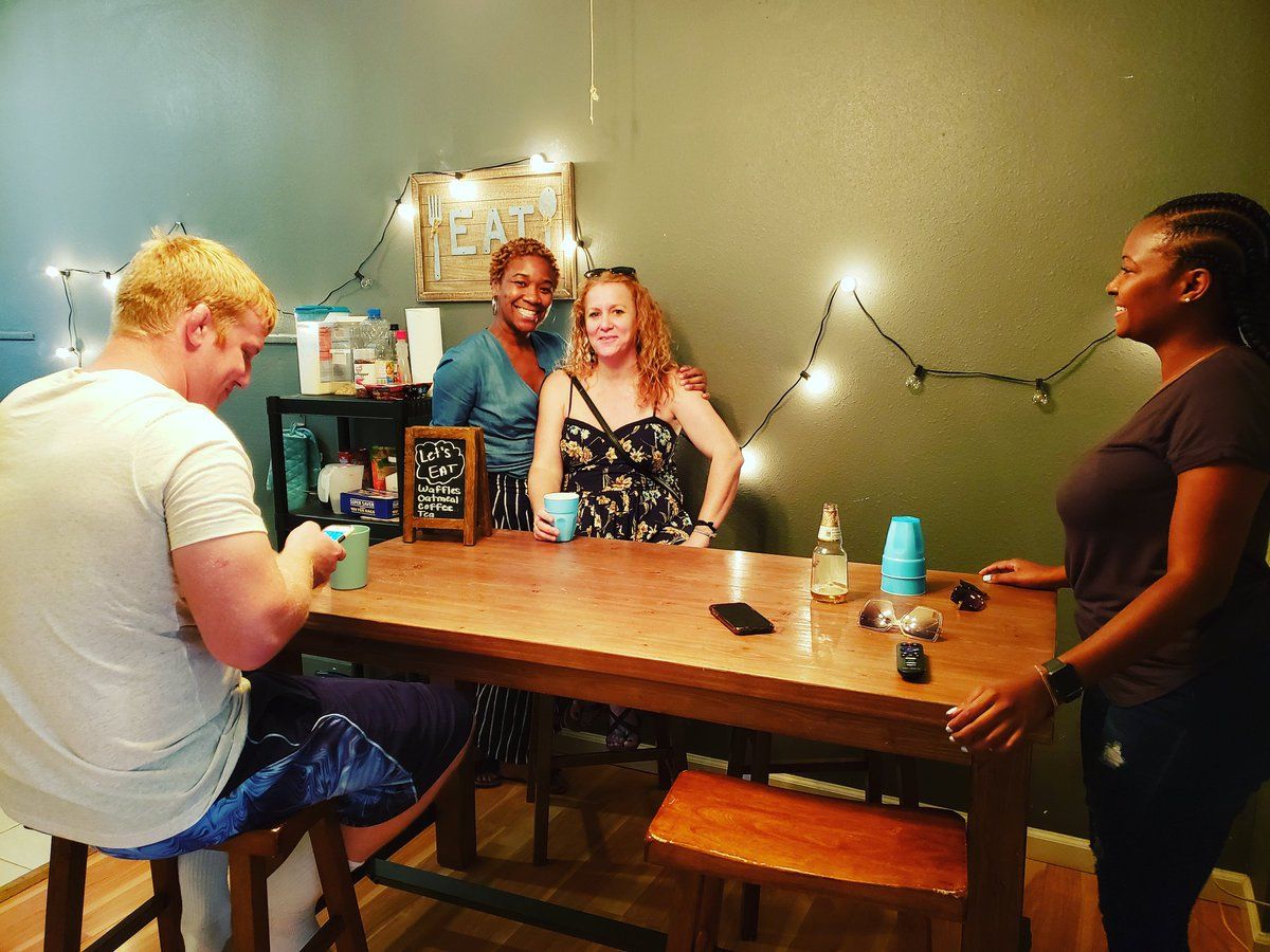 Four people gather around a wooden table in a room. String lights hang behind them.