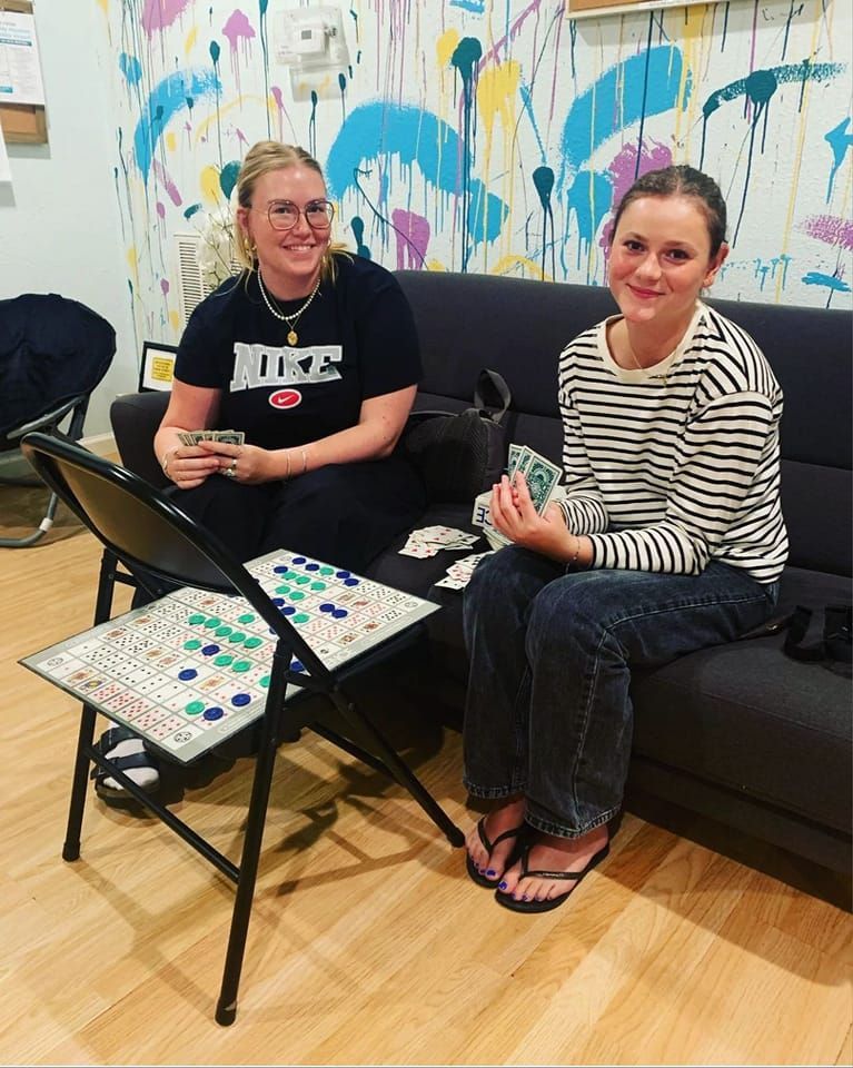 Two people playing a board game on a folding chair in front of a colorful wall.