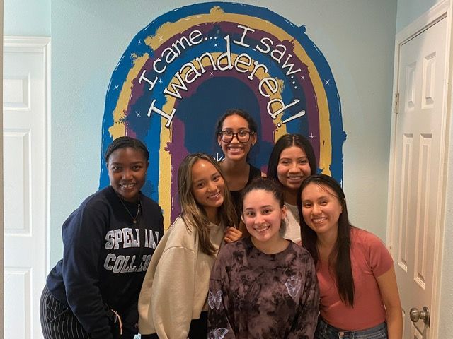 Six people smiling in front of a colorful wall mural that reads,