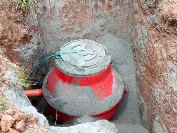 Red septic tank in a dirt trench, surrounded by sand, with a green lid and orange pipe.