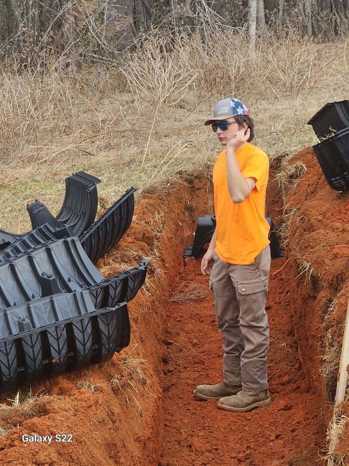 A person in an orange shirt and cap stands in a trench next to drainage infrastructure in a field.
