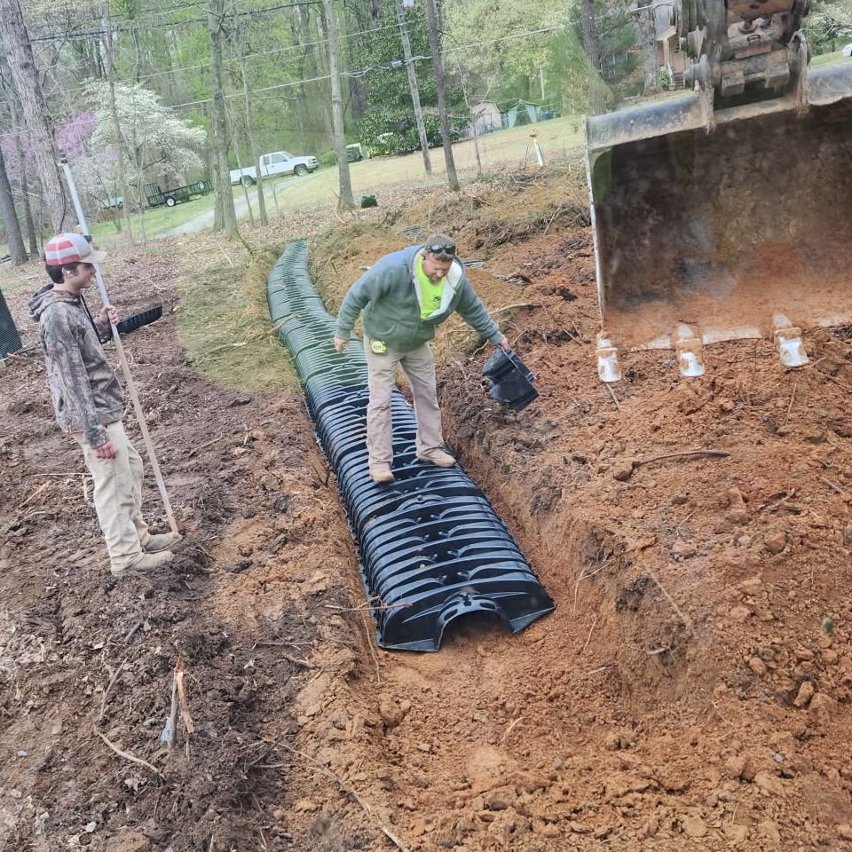 Two workers installing black drainage pipe in a trench; excavator in background.