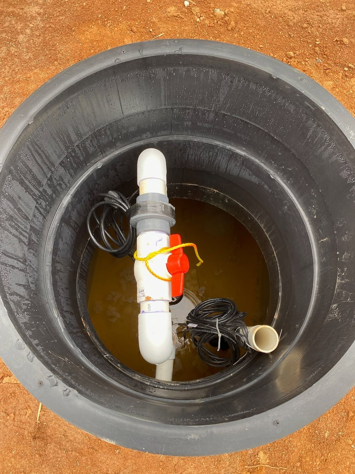 Black basin with water, PVC piping, valve, and wiring. Located outside on dirt.
