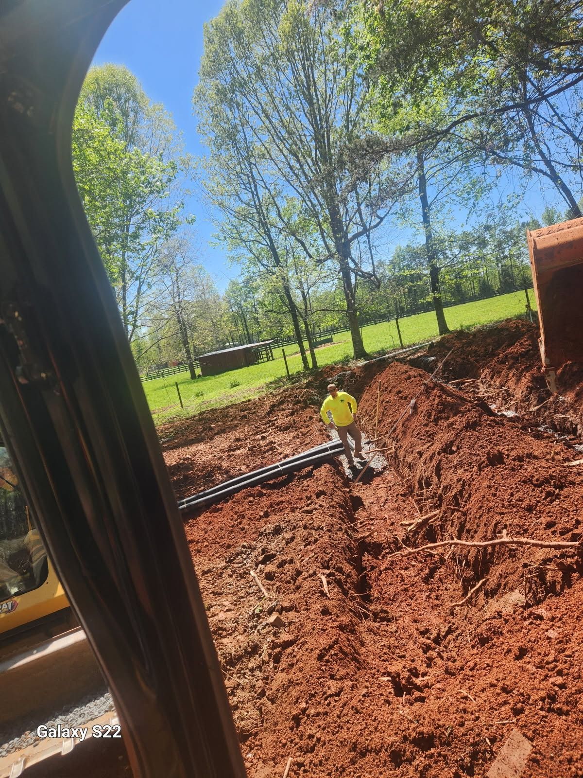 Black pipes laid in the trench; red dirt surrounds. Trees and a green field in the background.