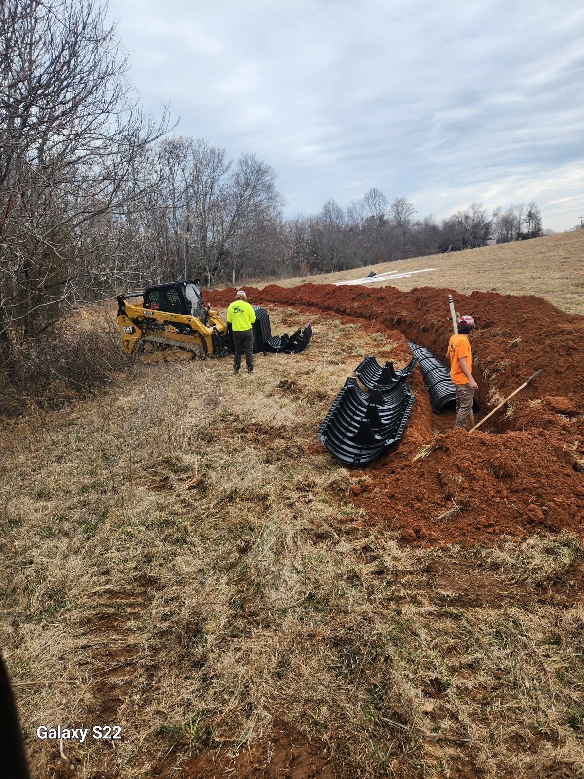 Construction site with workers, a backhoe, and drainage pipes in a field. Cloudy sky.