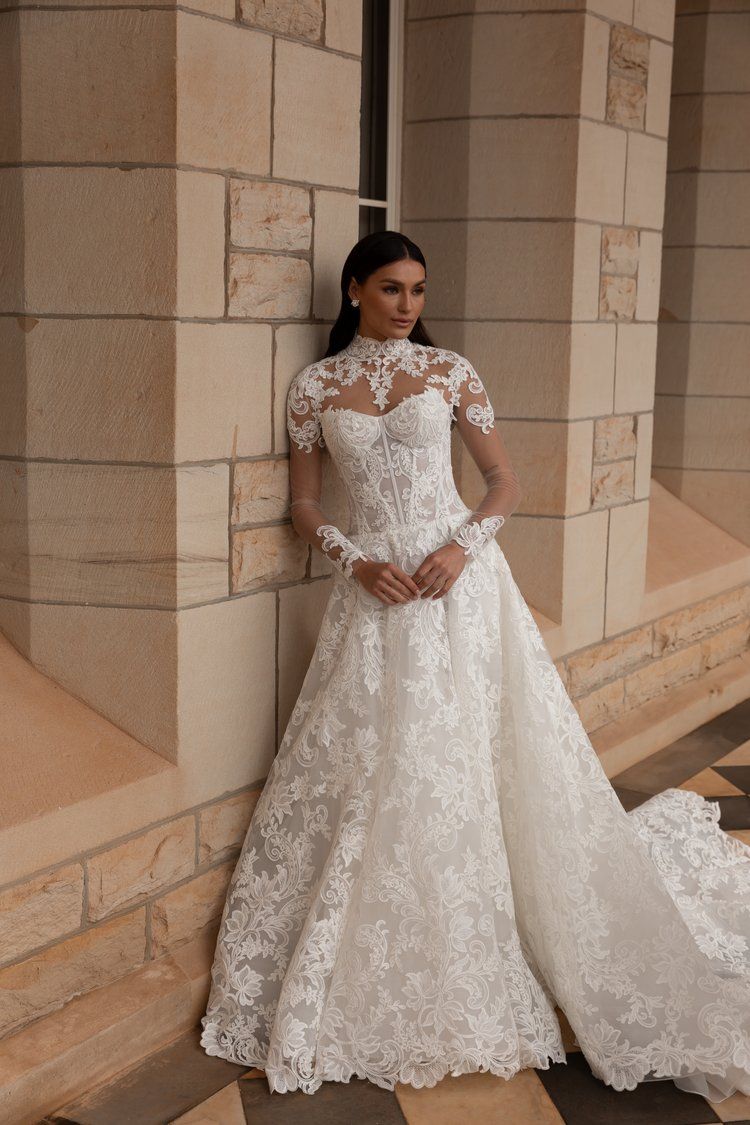 A woman in a white lace wedding dress is leaning against a brick wall.